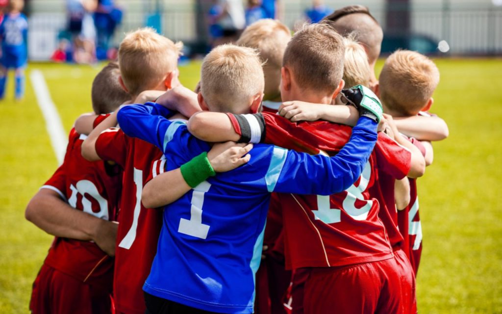 A group of young soccer players in red uniforms huddle together on a grassy field before a game.