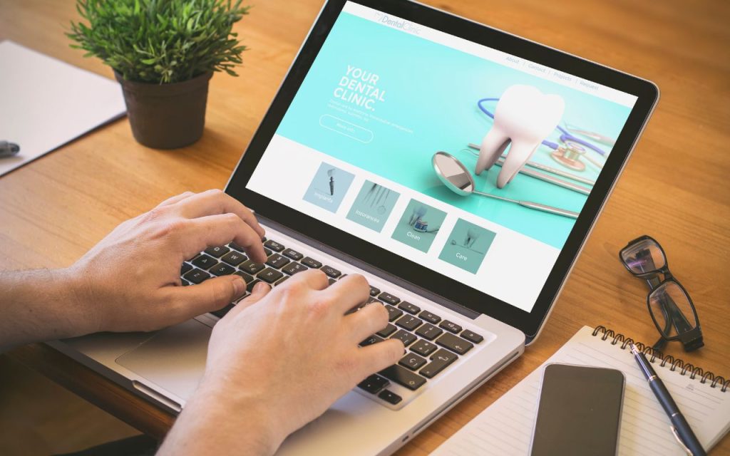 Person using a laptop displaying a dental clinic website, with a plant, glasses, notepad, and pen on a wooden desk.