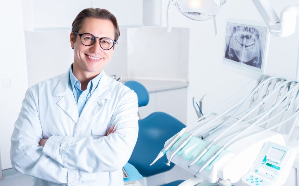 A dentist wearing glasses and a white coat stands smiling in a modern dental clinic, with dental equipment and an X-ray visible in the background.