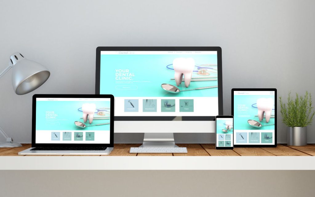 A desk with a desktop computer, laptop, tablet, and smartphone displaying the same dental clinic website, next to a lamp and small plant.