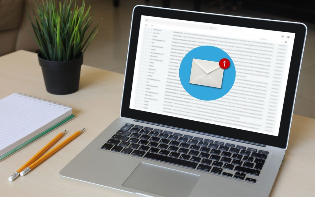 AI-driven email marketing that practically writes itself. A laptop displaying an email inbox with a notification for one new message; a notebook, two pencils, and a potted plant are on the desk.