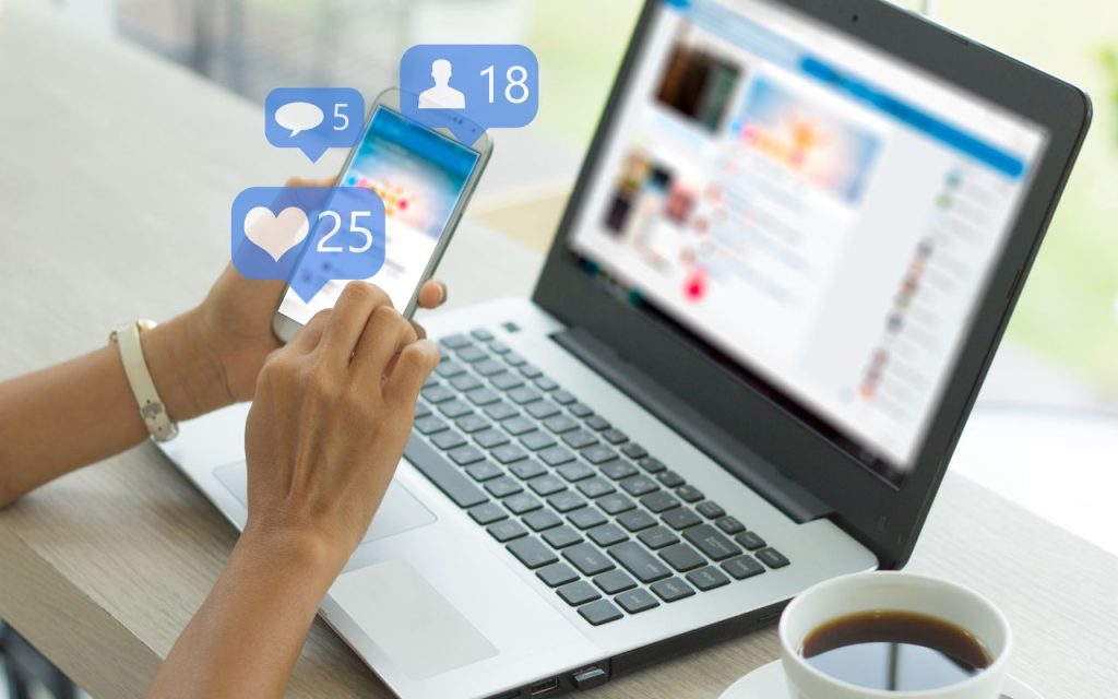 AI-enhanced social media scheduling to maximize engagement. A person uses a smartphone and a laptop at a desk with a cup of coffee. Social media notification icons for likes, messages, and followers appear on the phone.