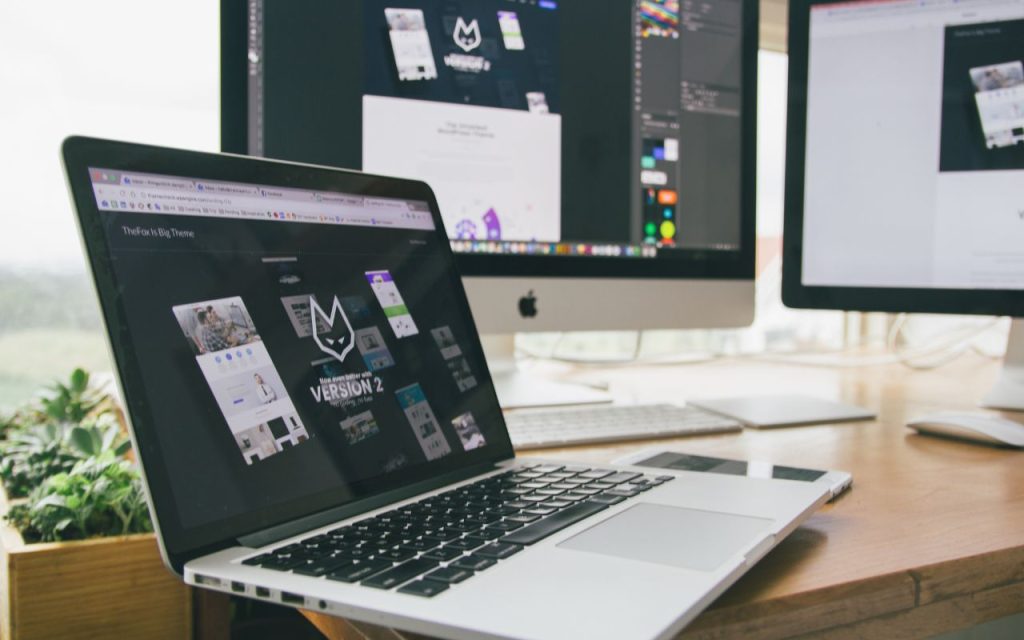 AI-powered design tools for professional marketing materials in minutes. A laptop and two desktop monitors display web design software and website mockups on a wooden desk near a window, with a small plant nearby.