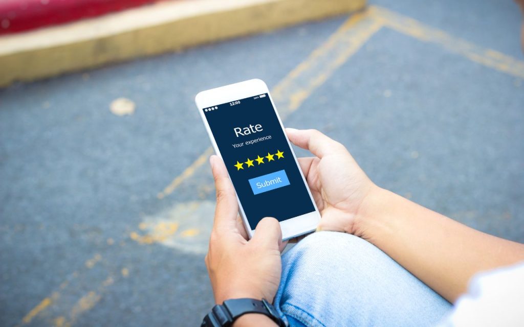 Use AI for automated review requests to build trust and credibility. A person holds a smartphone displaying a five-star rating form on a "Rate your experience" screen, with a blue "Submit" button, while sitting outdoors on pavement.