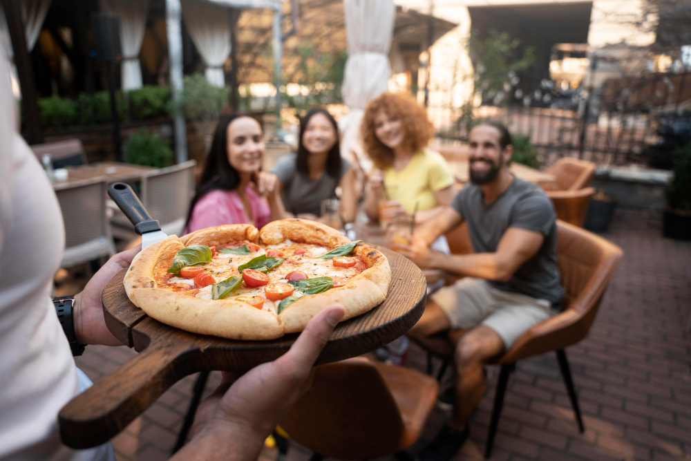 A person serves a pizza on a wooden tray to four people sitting at an outdoor restaurant table.