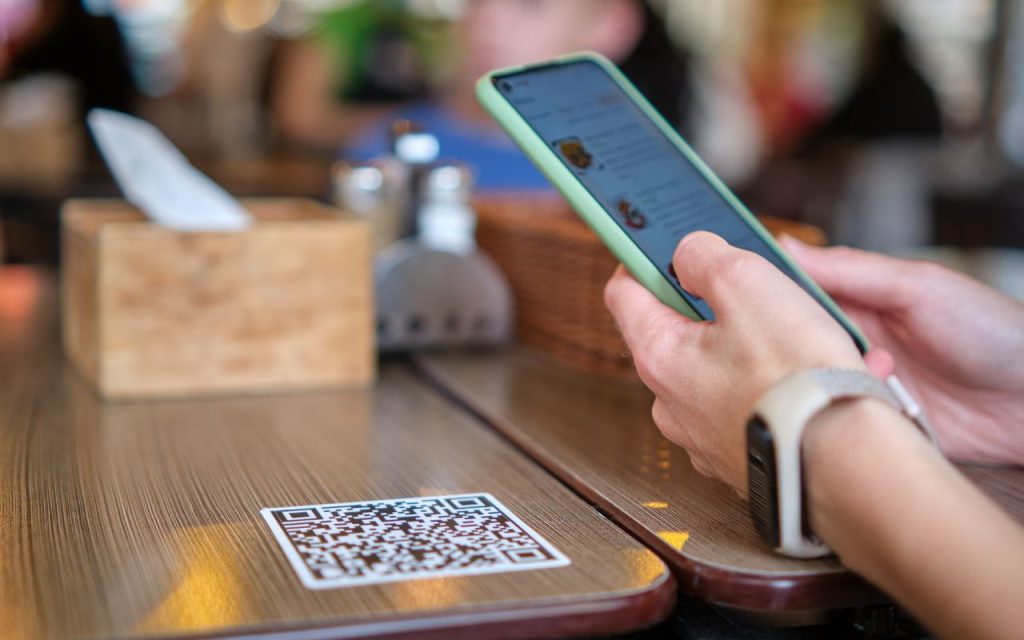A person holding a smartphone scans a QR code on a wooden table at a restaurant; a tissue box and condiments are also on the table.