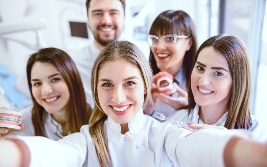 Five young adults in white coats smile for a group photo in a dental office; two hold dental models and one holds a toothbrush.